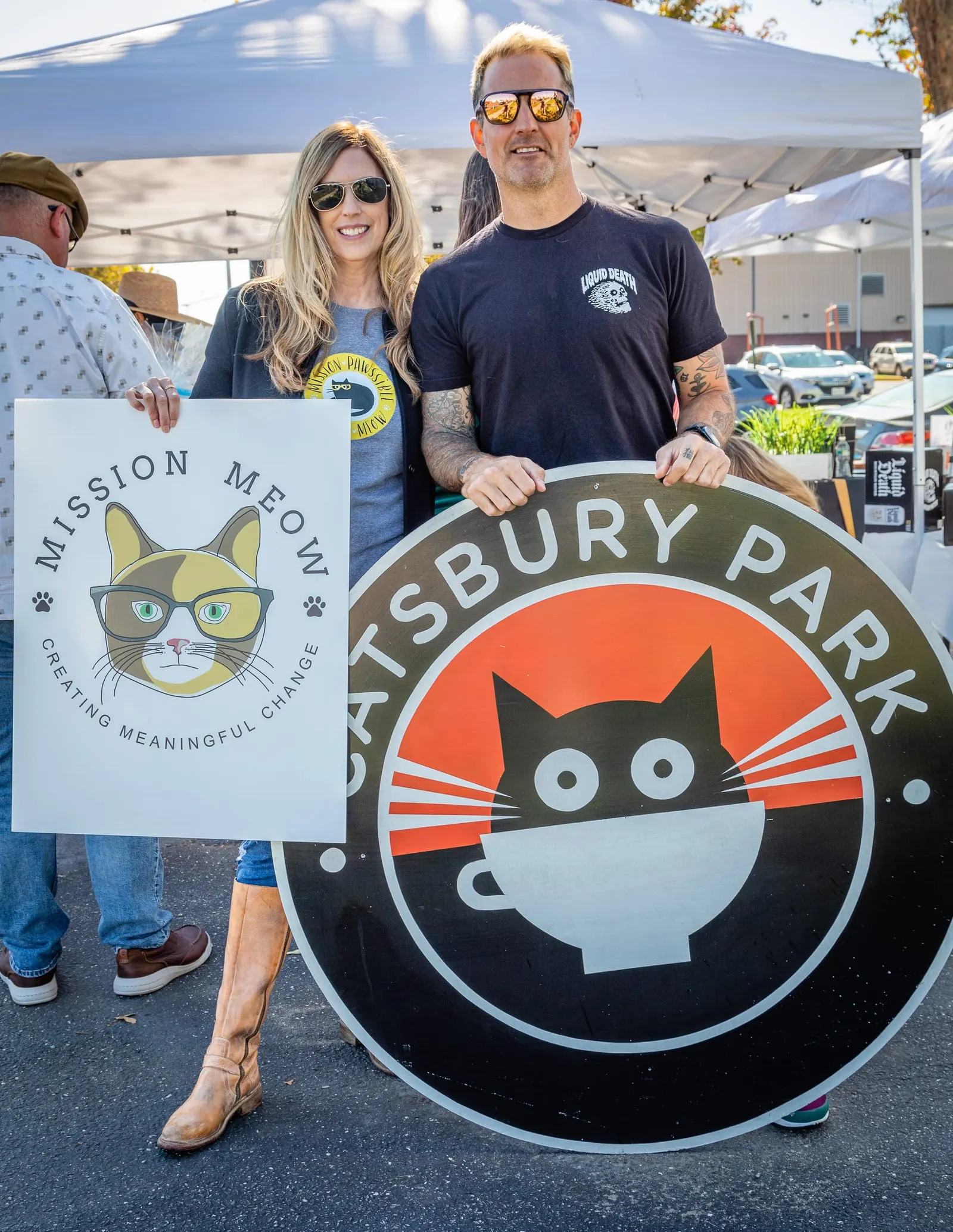 Sally Williams holds a Mission Meow sign beside a man holding a large round Catsbury Park sign at an outdoor event