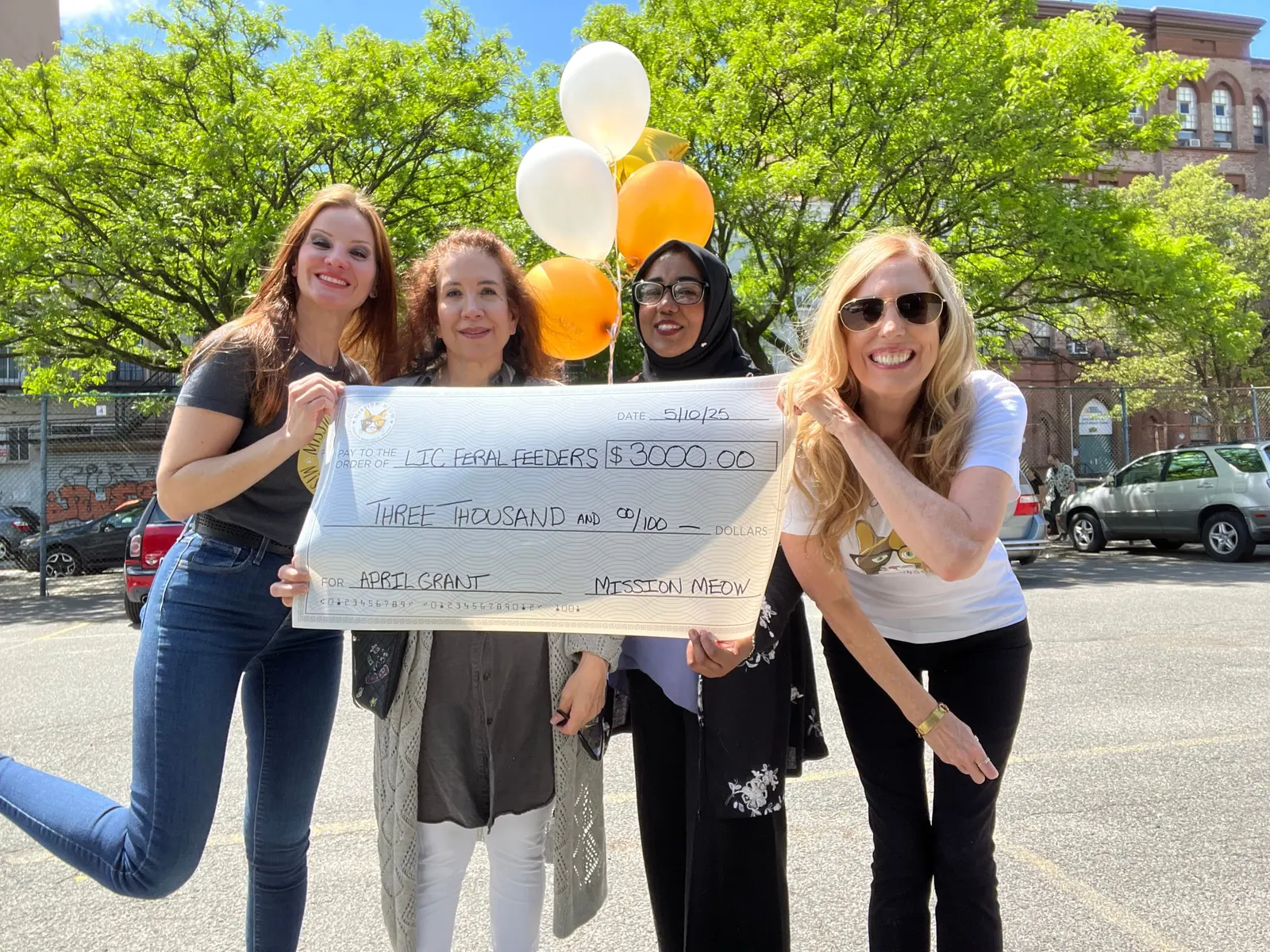 Four women jump and cheer outdoors holding a giant Mission Meow check, with balloons