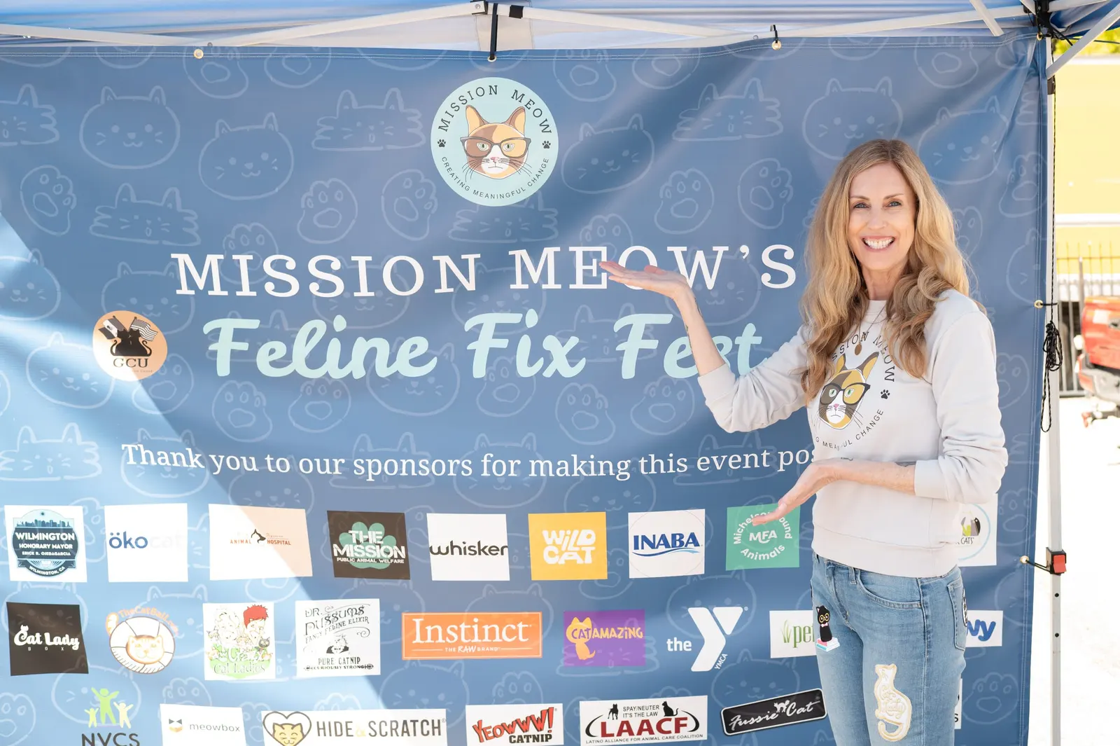 Sally Williams in front of Mission Meow’s Feline Fix Fest banner, which thanks the event’s sponsors with their logos