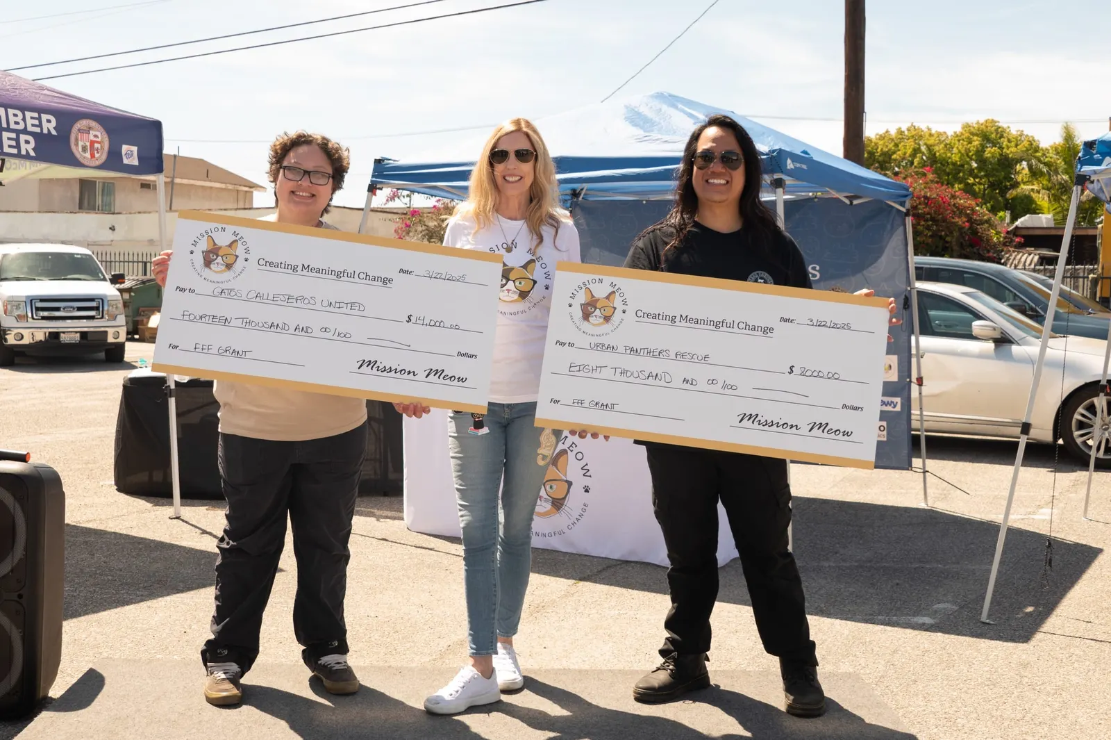 Two rescue leaders hold giant Mission Meow checks for $14,000 to Gatos Callejeros United and $8,000 to Urban Panthers Rescue, with a woman in a Mission Meow T-shirt between them