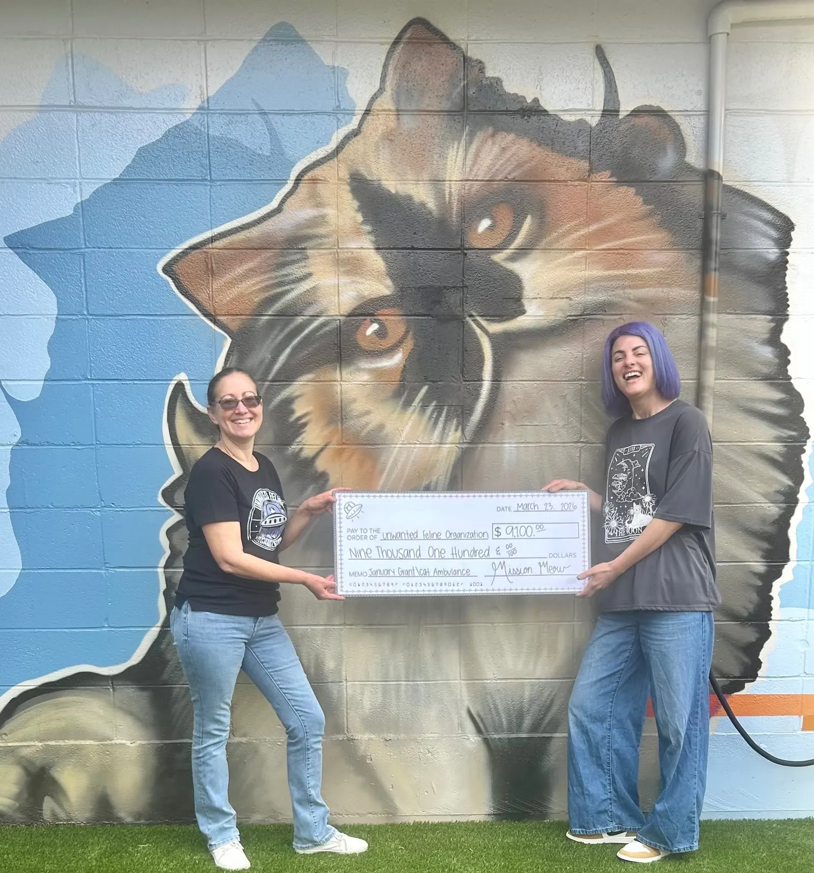 Two women hold a giant Mission Meow check made out to Unwanted Feline Organization in front of a large painted cat mural
