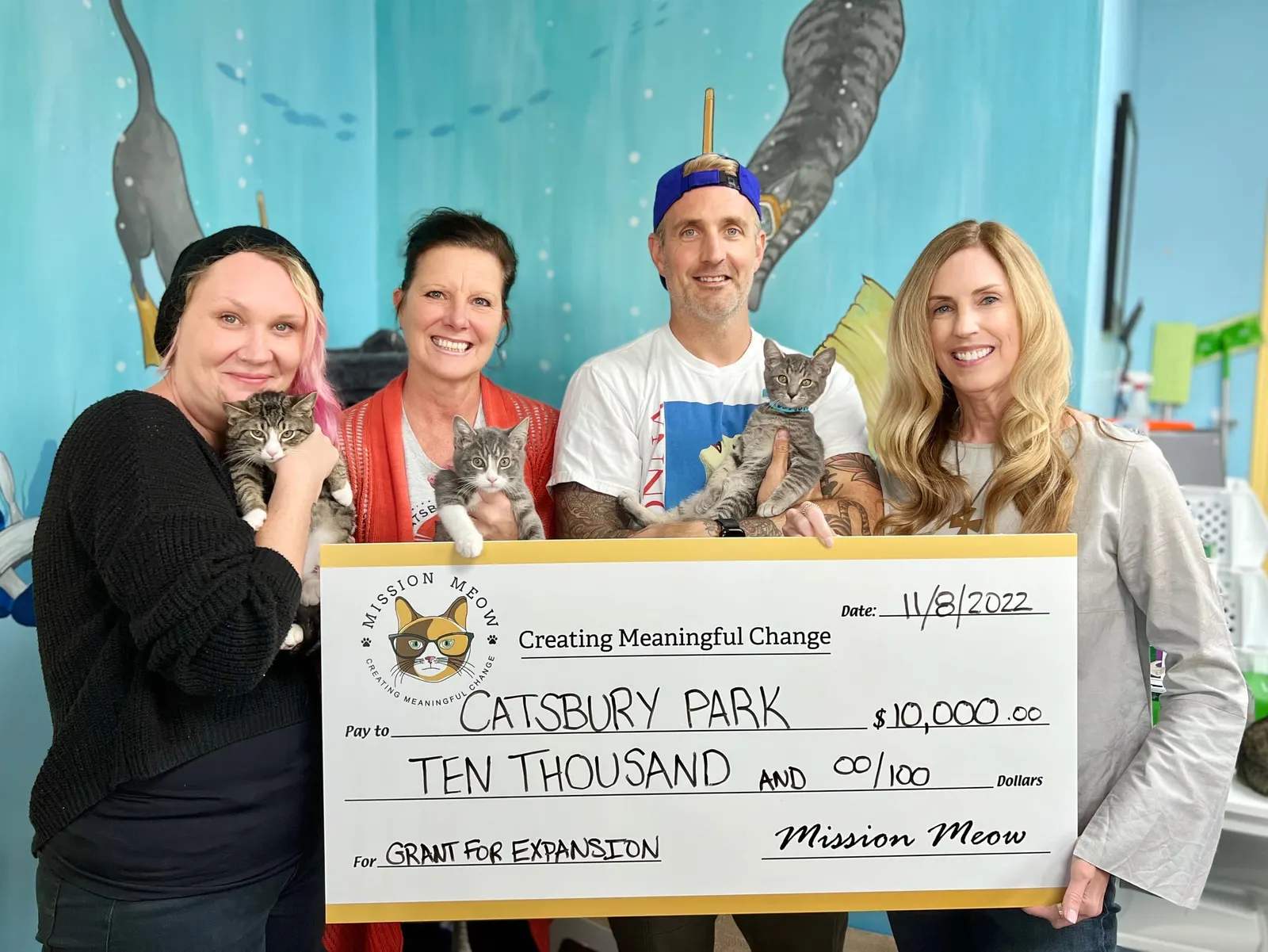 Four Catsbury Park team members hold kittens and a $10,000 Mission Meow check in front of an aqua cat mural