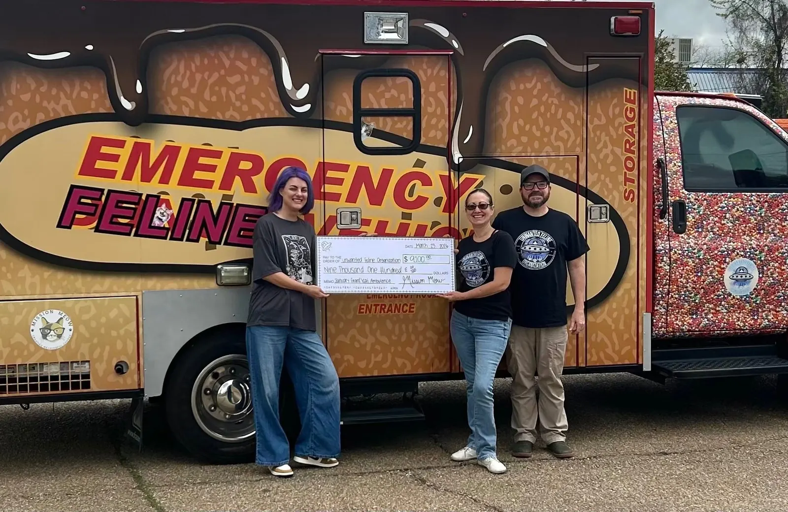 Three people hold a giant Mission Meow check made out to Unwanted Feline Organization in front of an ambulance painted with “Emergency Feline” lettering